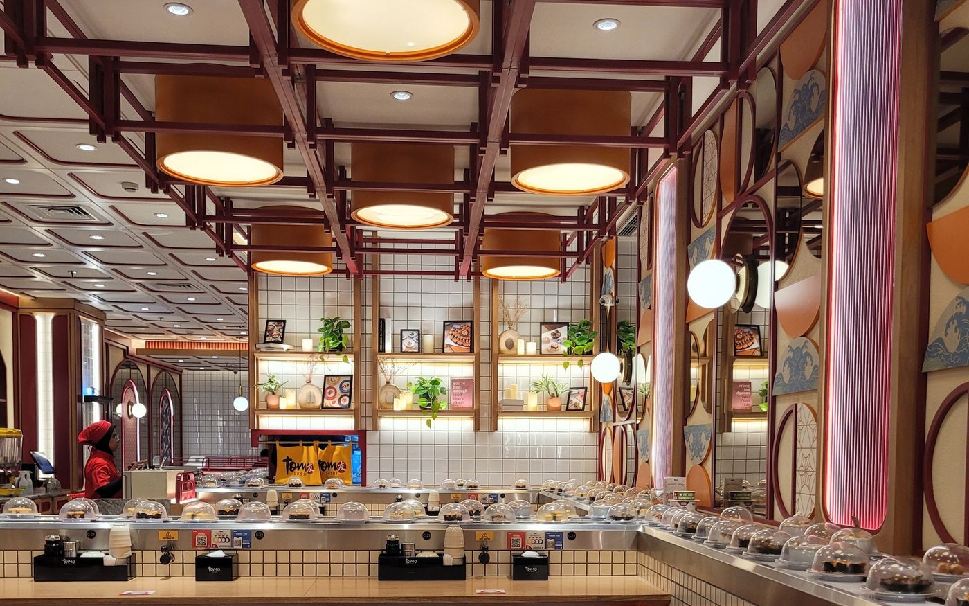 The counter and conveyor belt of a kaiten sushi restaurant, plates under domed covers, a hot-water tap and a teacup at the seat