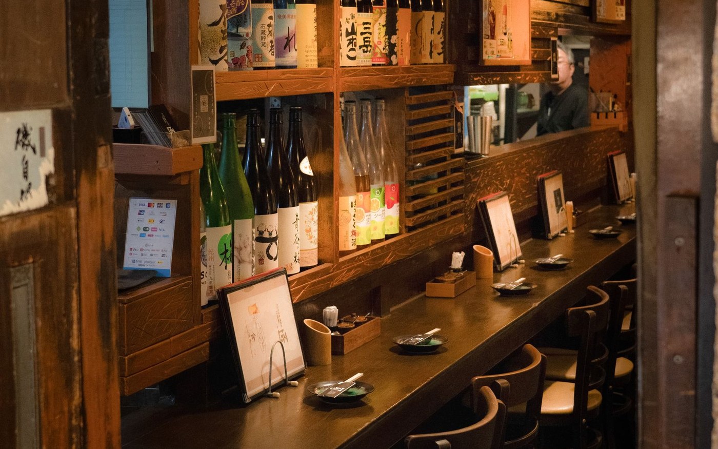 A sushi counter in a small Japanese restaurant: a wooden bar, place settings, menus and a shelf of sake bottles