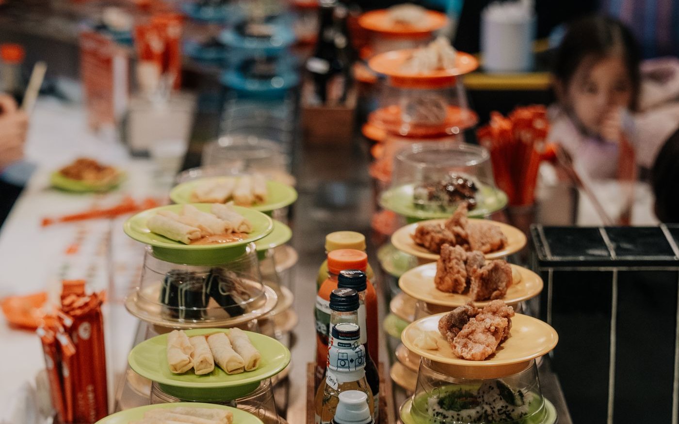 Plates of sushi under clear plastic domes on a conveyor belt, a diner reaching for one plate