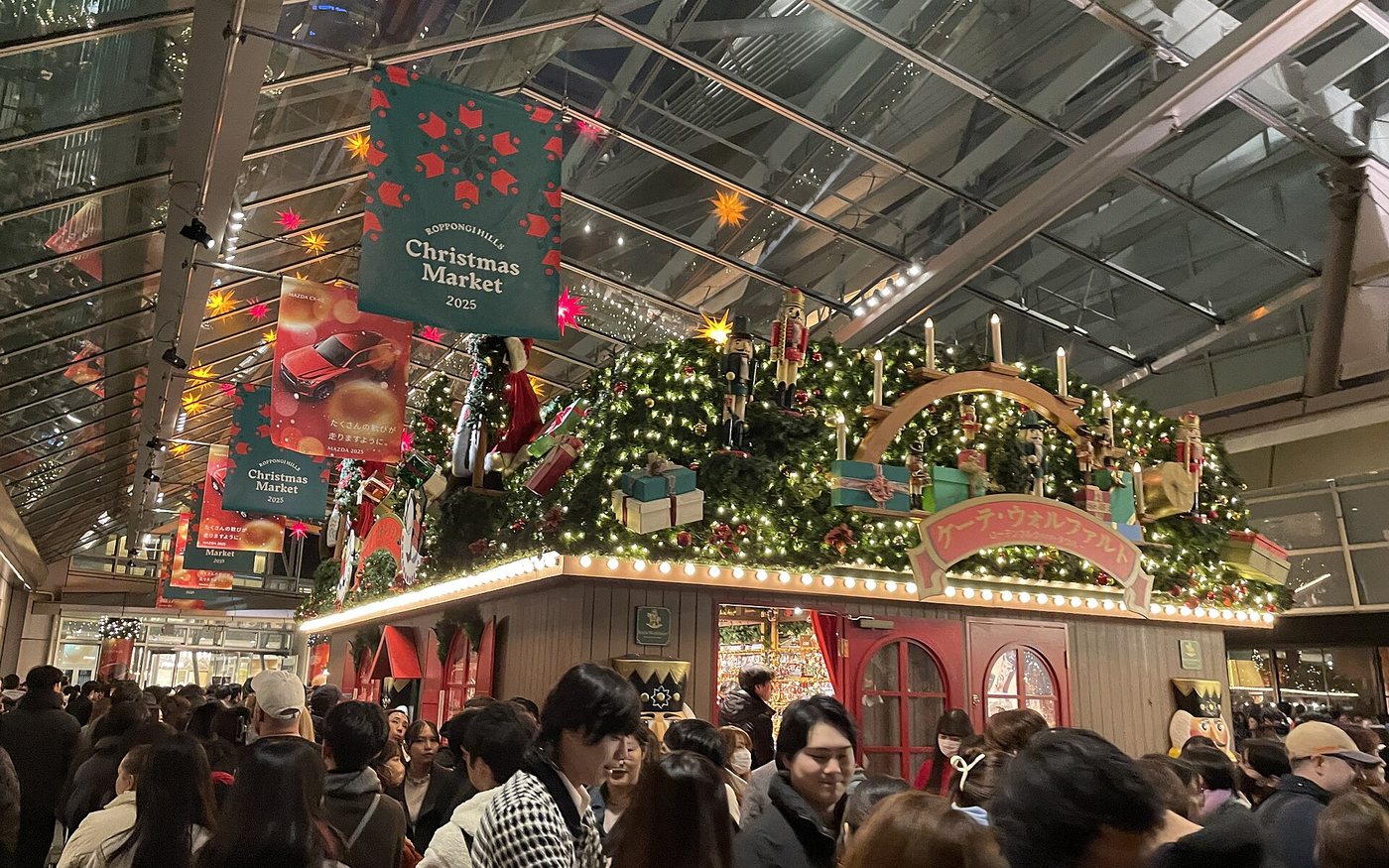 Crowds in winter coats in front of a wooden Christmas market hut hung with garland, under a glass roof