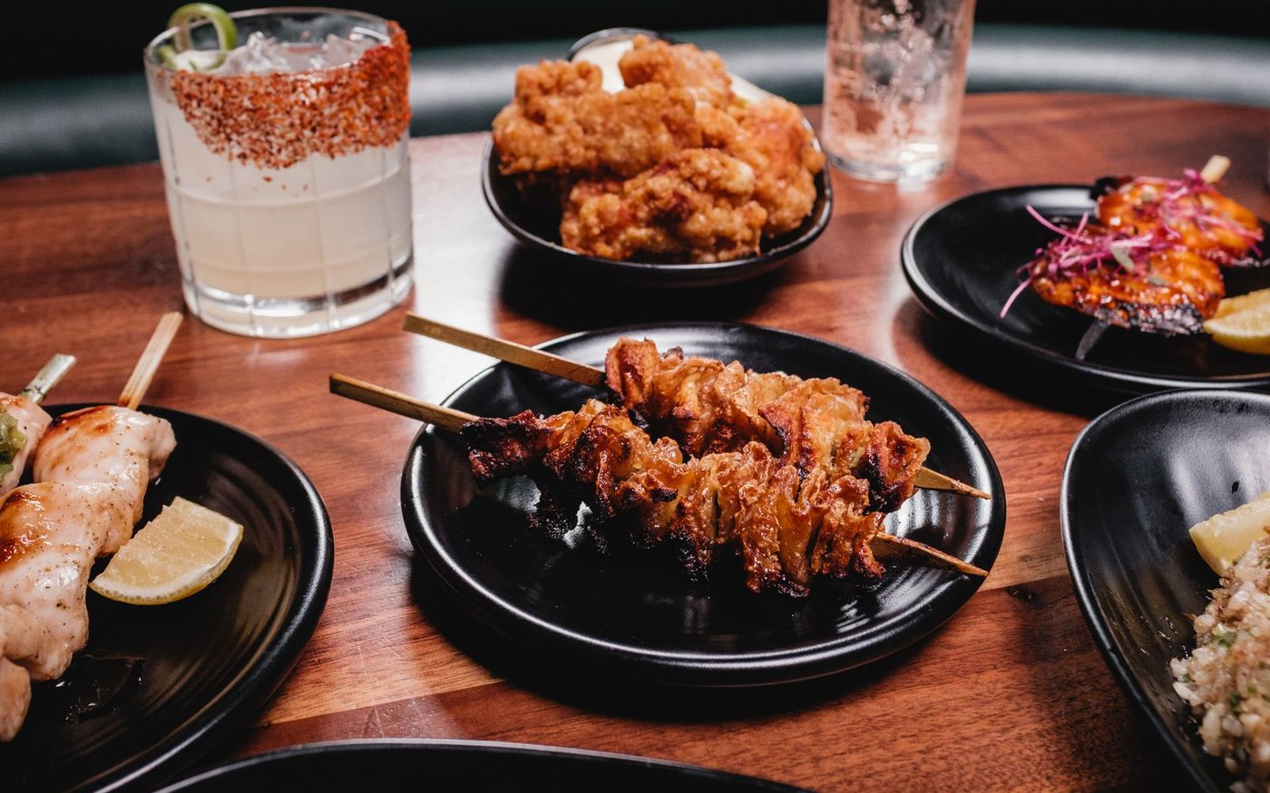 An izakaya table of grilled chicken skewers on small black plates with a tall iced drink