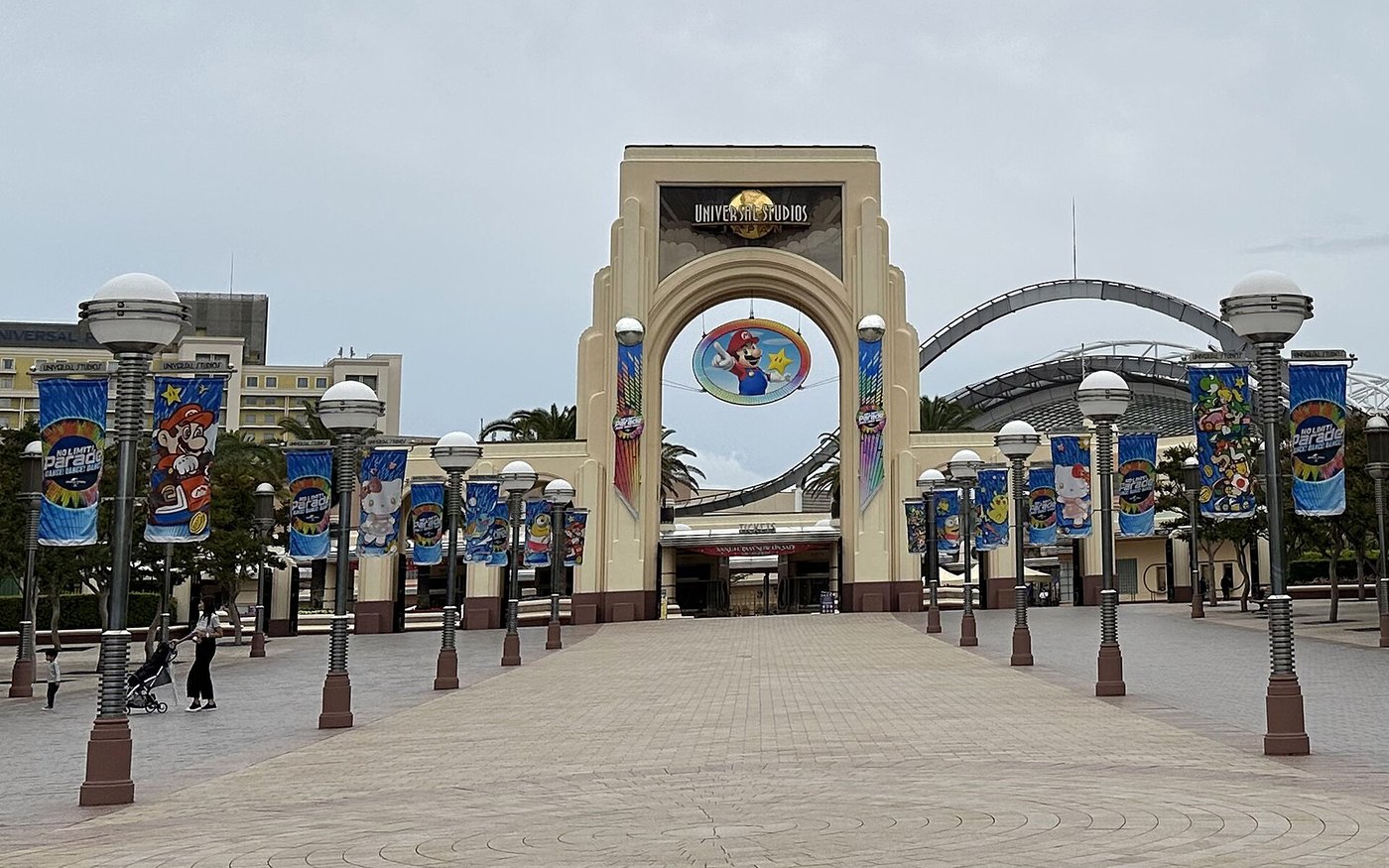 The Universal Studios Japan entrance arch with a globe logo, Mario graphic and NO LIMIT! Parade banners on both sides of an empty forecourt