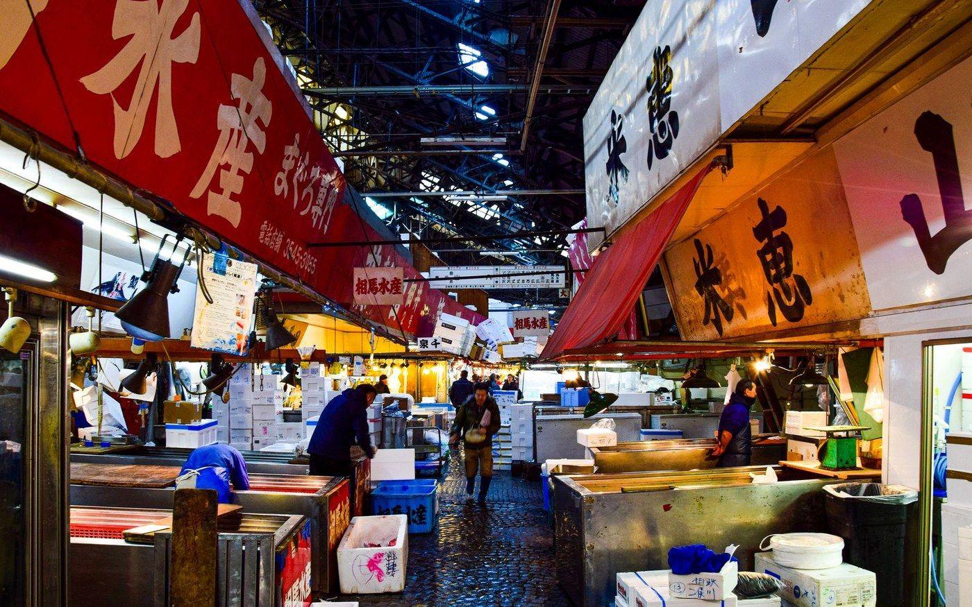 A covered market street in Japan with red banners, food stalls and shoppers