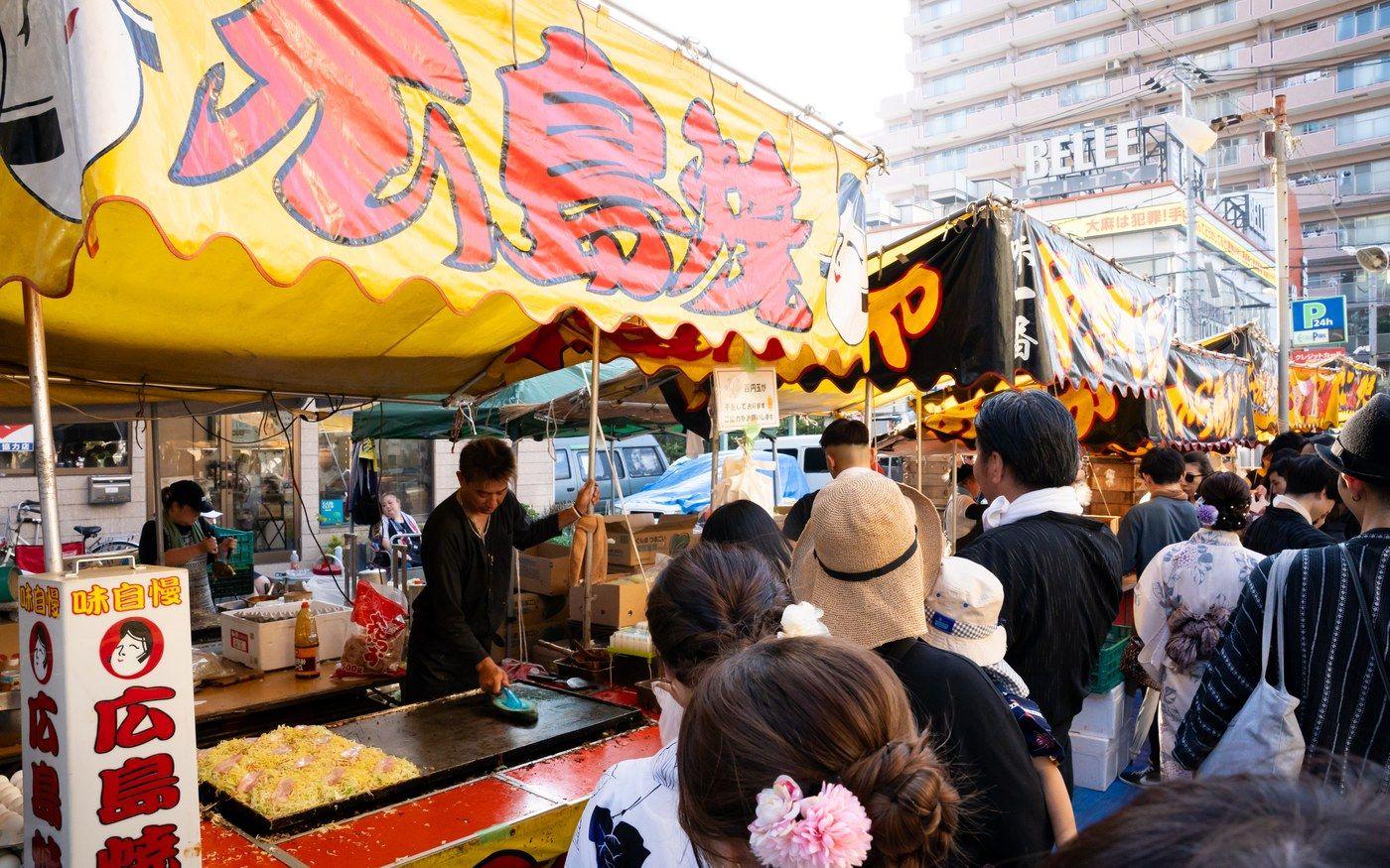 A festival food stall cooking Hiroshima-style okonomiyaki on a griddle, with customers in yukata outside