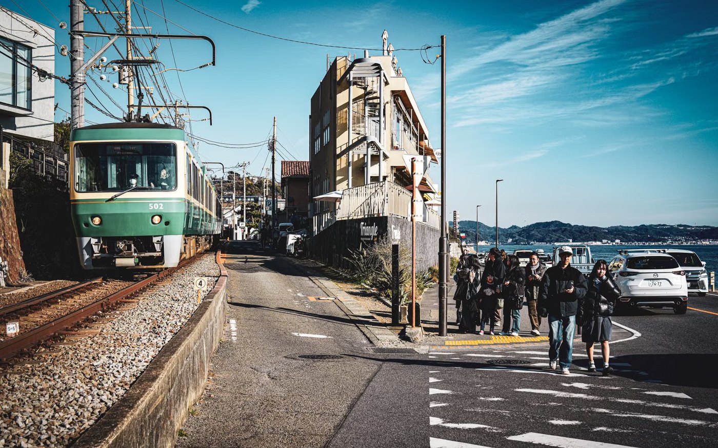 A green Enoden tram running along the street with pedestrians and overhead wires