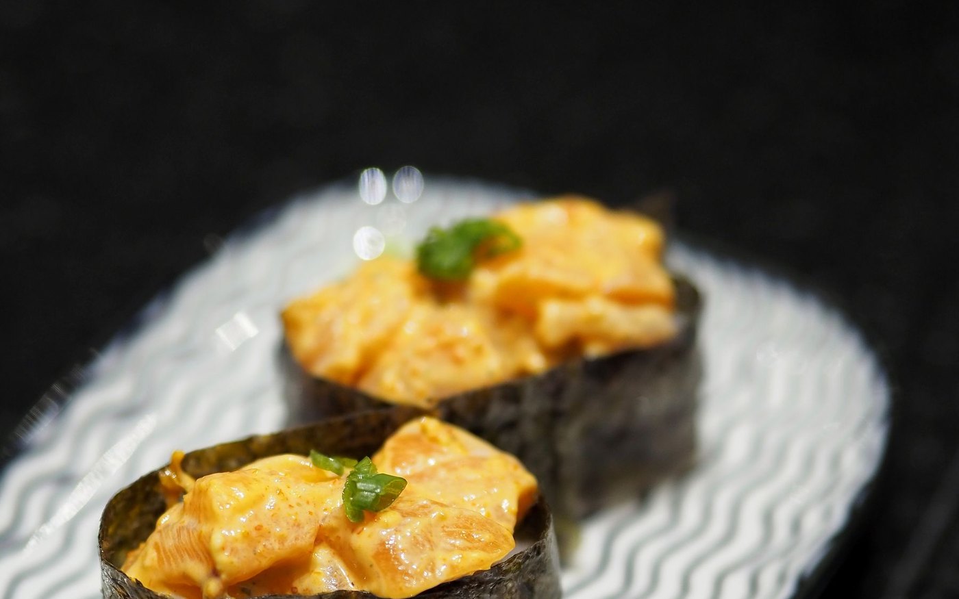 Two gunkan maki sushi with a creamy orange topping and a herb garnish on a ridged white plate