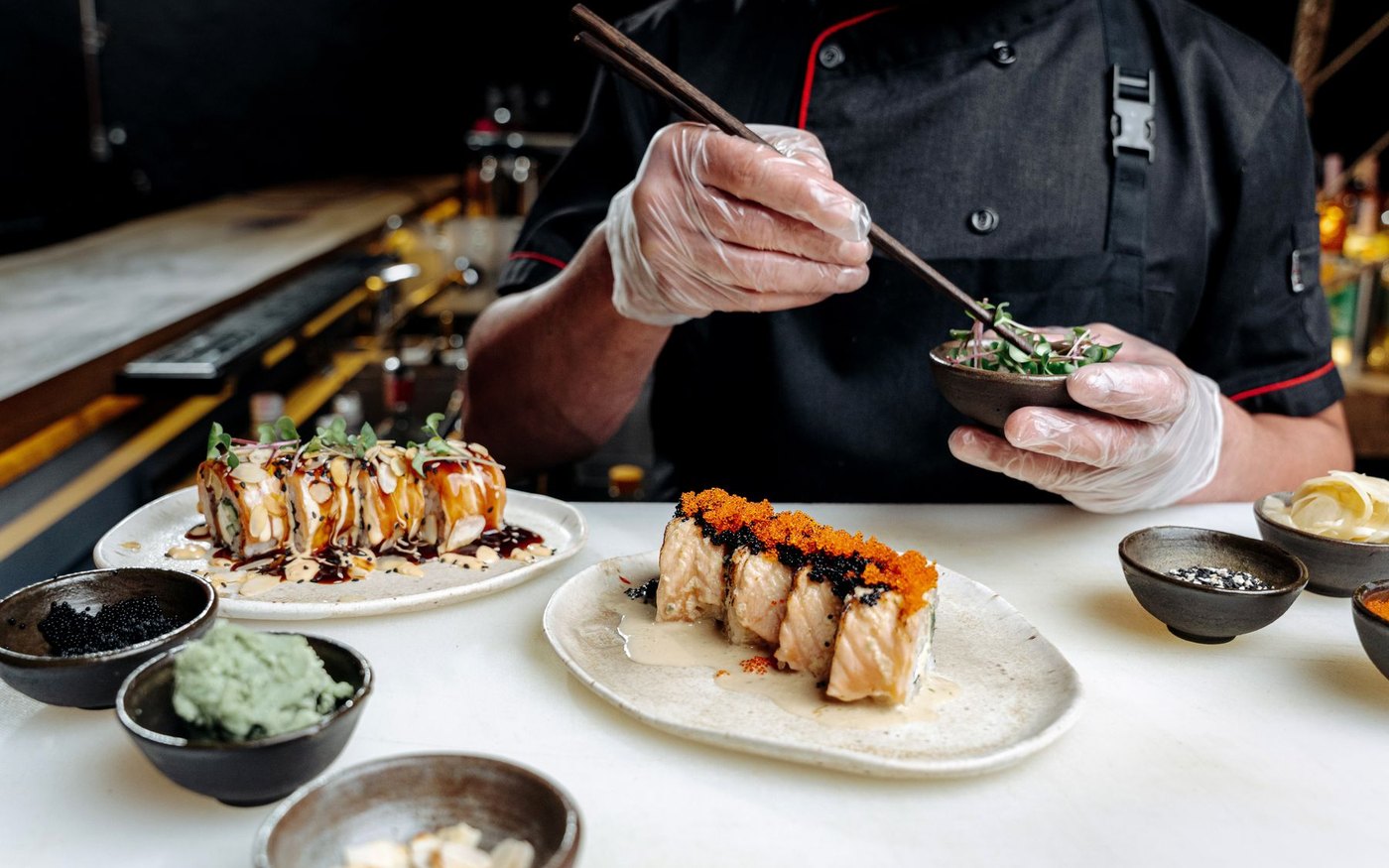 A chef in black and white gloves using chopsticks to garnish a sushi roll at a restaurant counter, plates laid out in front