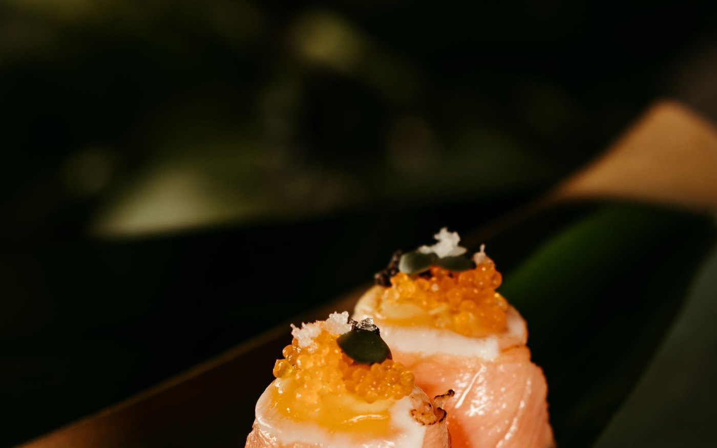 Two pieces of salmon nigiri topped with roe and herbs on a dark dish, shot close up