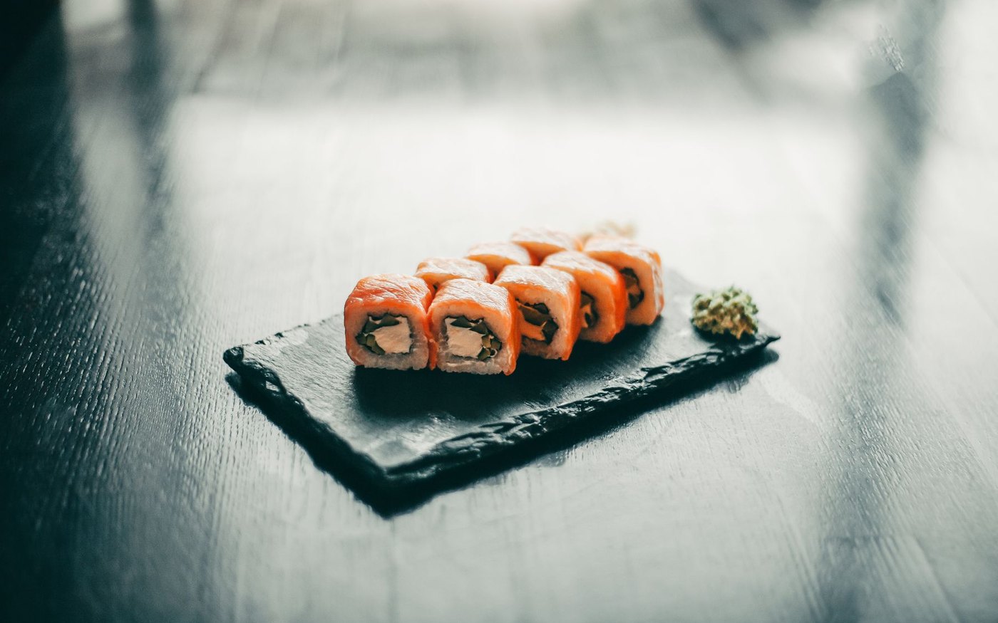 Five cut sushi rolls with salmon, avocado and cucumber on a dark slate plate beside a mound of green wasabi