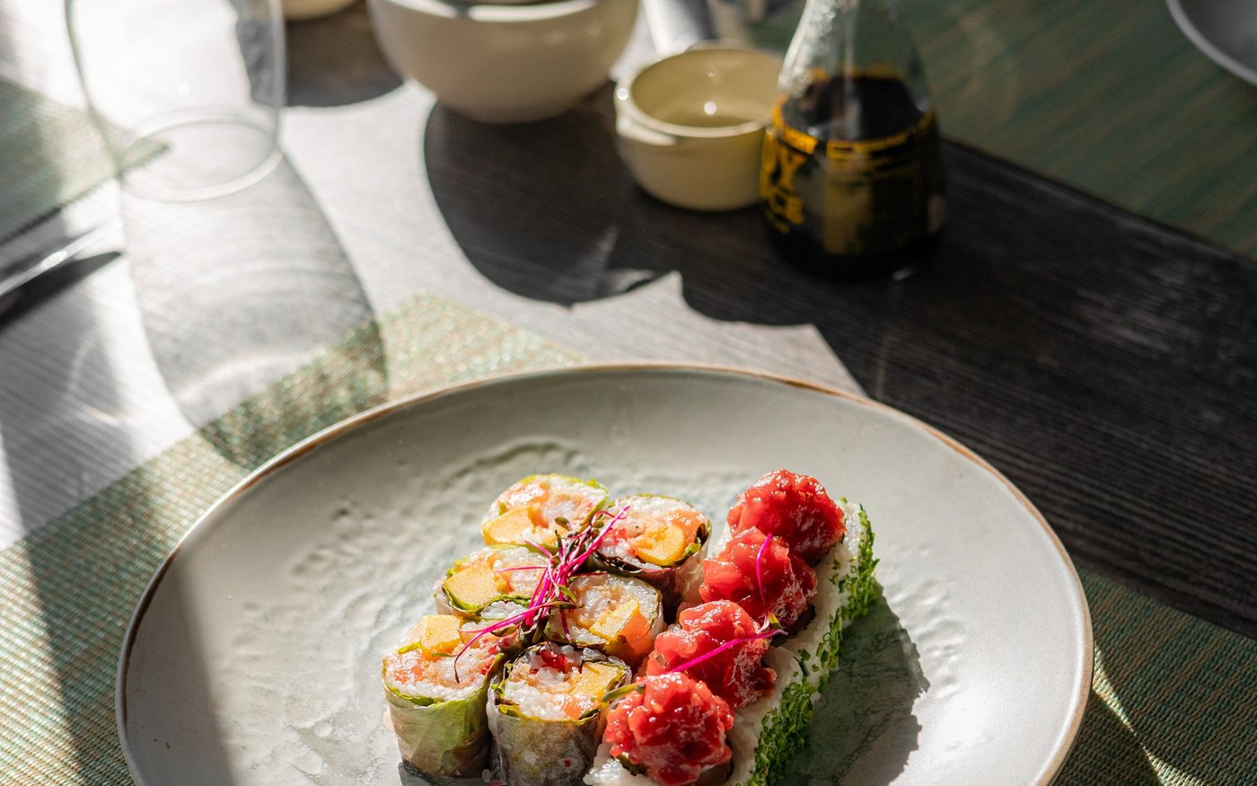 A plate of sushi rolls topped with red roe on a speckled plate, with a soy sauce bottle and a glass on a sunlit table