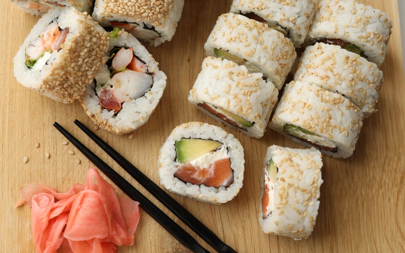 Sushi rolls on a light wooden board with a pair of black chopsticks and a pile of pink pickled ginger