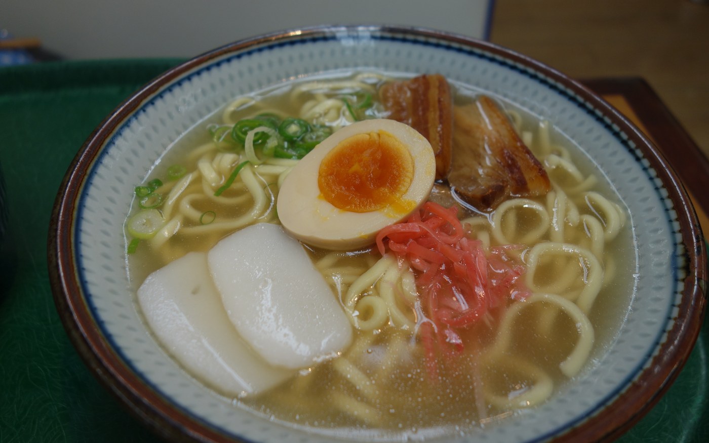 A bowl of Okinawa soba with thick wheat noodles, braised pork, fish cake, pickled ginger and scallion in a light broth