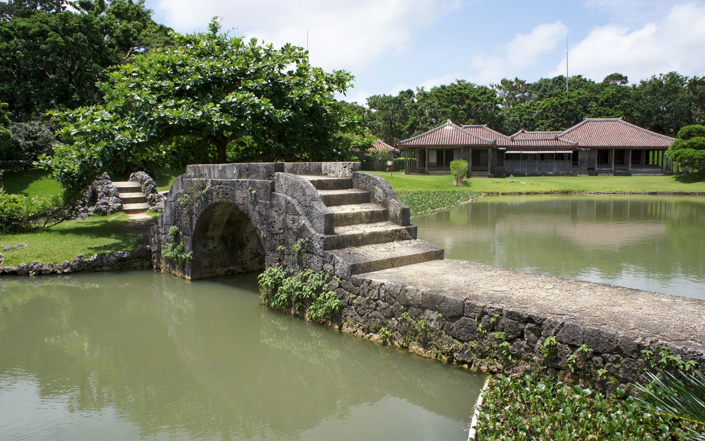 A stone bridge over the pond at Shikina-en, with trees and a tiled-roof pavilion behind it
