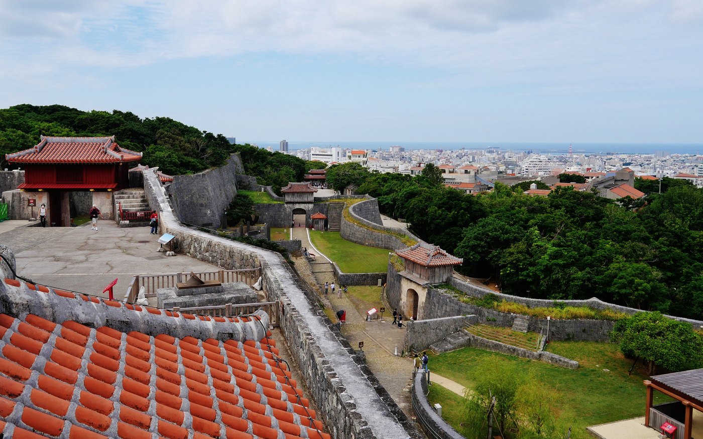 Shuri Castle's stone walls with red-tiled roofs behind them and the city of Naha in the distance