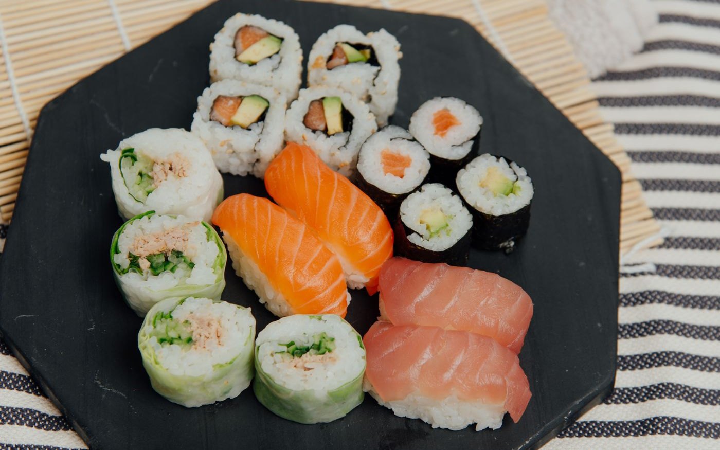An assortment of nigiri and rolls on a black plate, salmon and tuna pieces beside a bamboo mat