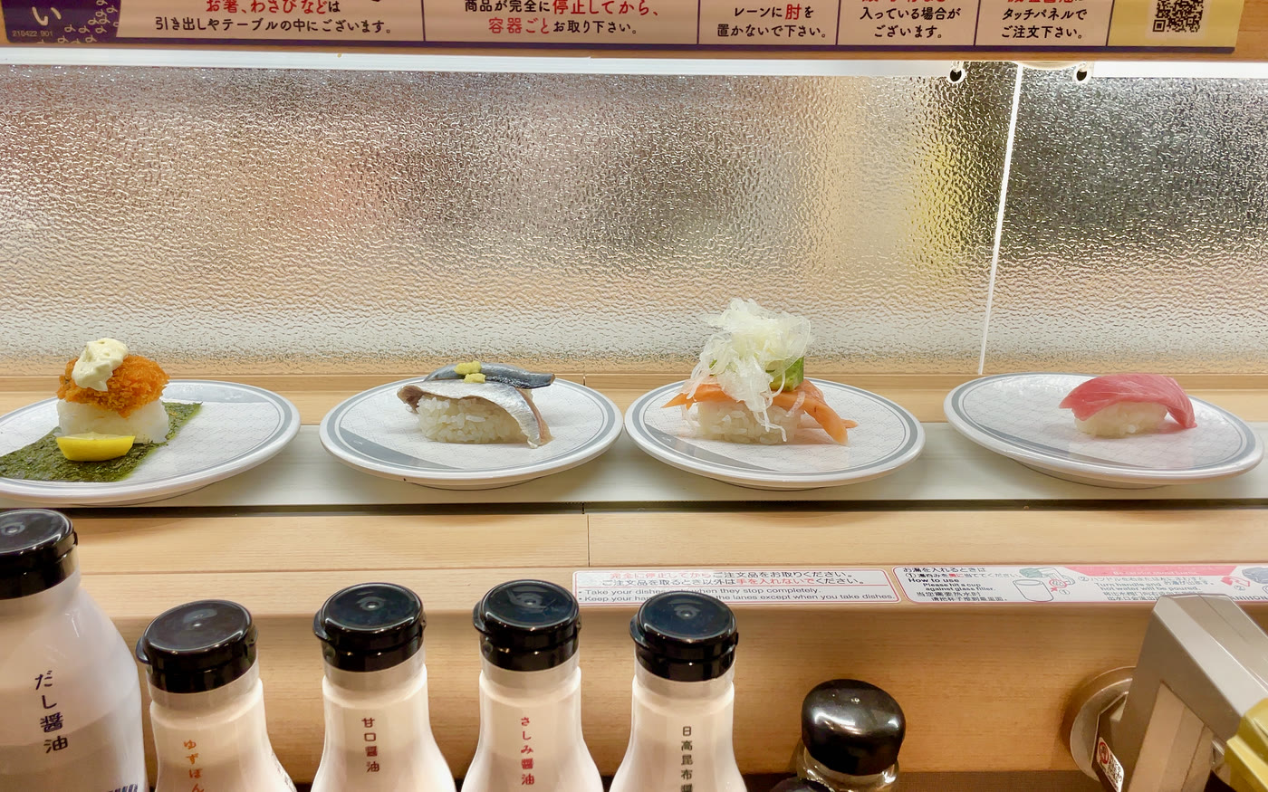 Four plates of sushi travelling on the conveyor belt of a kaiten sushi bar, soy sauce bottles on the counter in front
