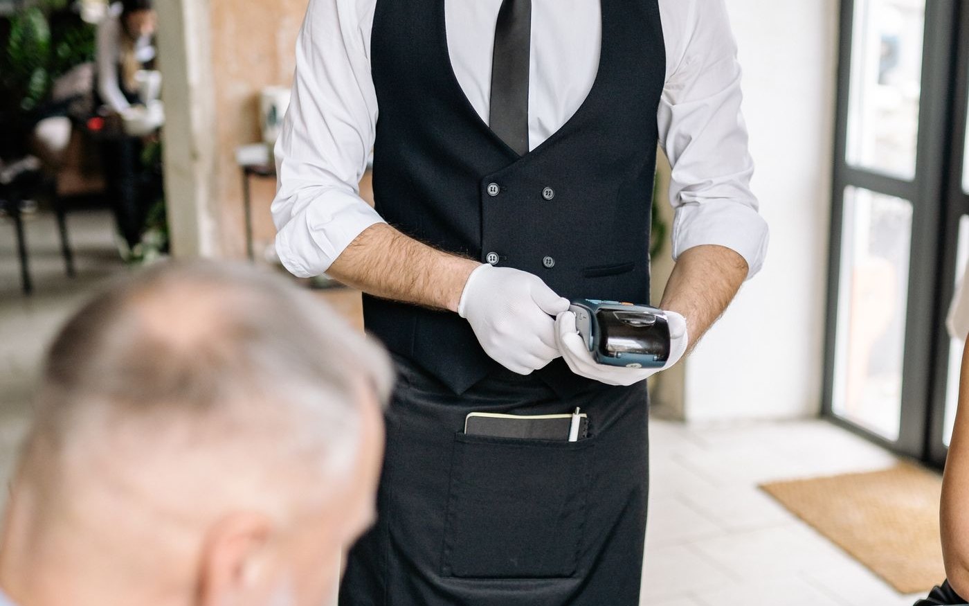 A waiter holding a card payment terminal at a restaurant table while a customer holds the bill