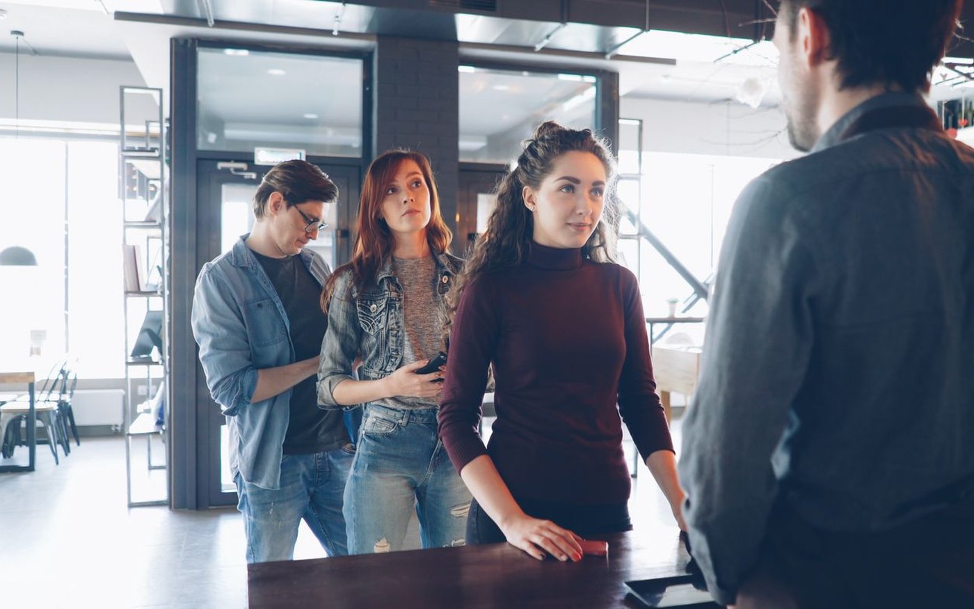 Three customers waiting at a restaurant counter, one of them looking at a phone