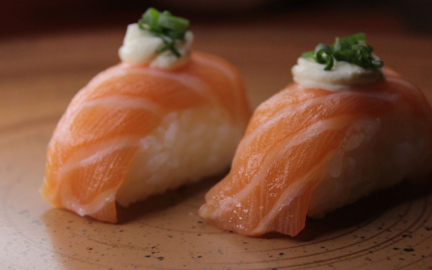Two pieces of salmon nigiri with a creamy topping and chopped spring onion on a speckled plate
