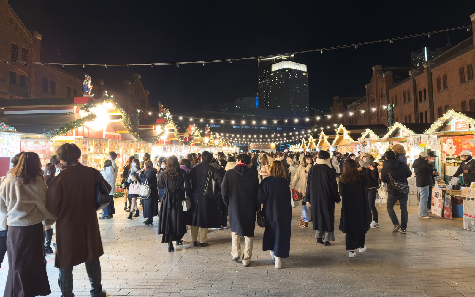 The Christmas market at Yokohama Red Brick Warehouse at night: wooden huts, crowds and a lit tree between the brick warehouses