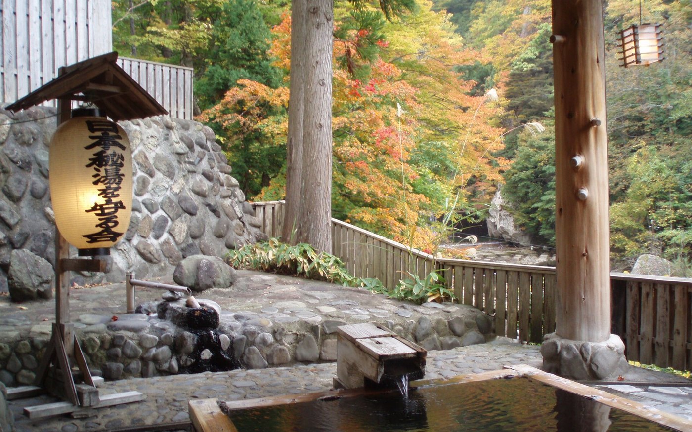 An outdoor hot-spring bath edged in rock, autumn trees behind it