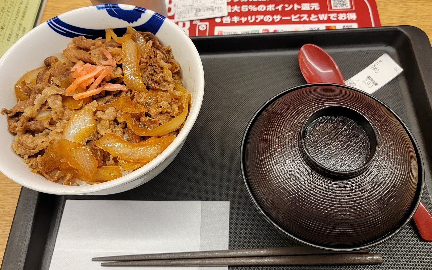 A large Matsuya beef bowl on a tray with a lidded bowl of soup, chopsticks, pickled ginger and a receipt