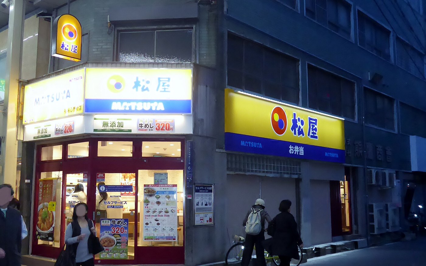 A Matsuya branch lit up at night on a city street, the sign above the glazed frontage