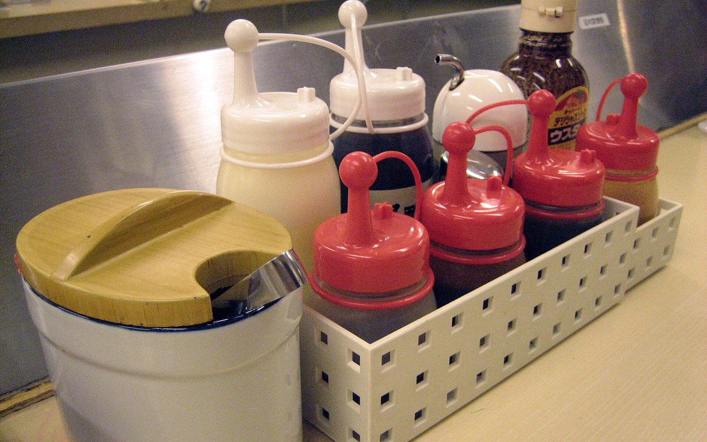 The condiment caddy on a Matsuya counter, sauce bottles with red caps and a yellow-lidded dish