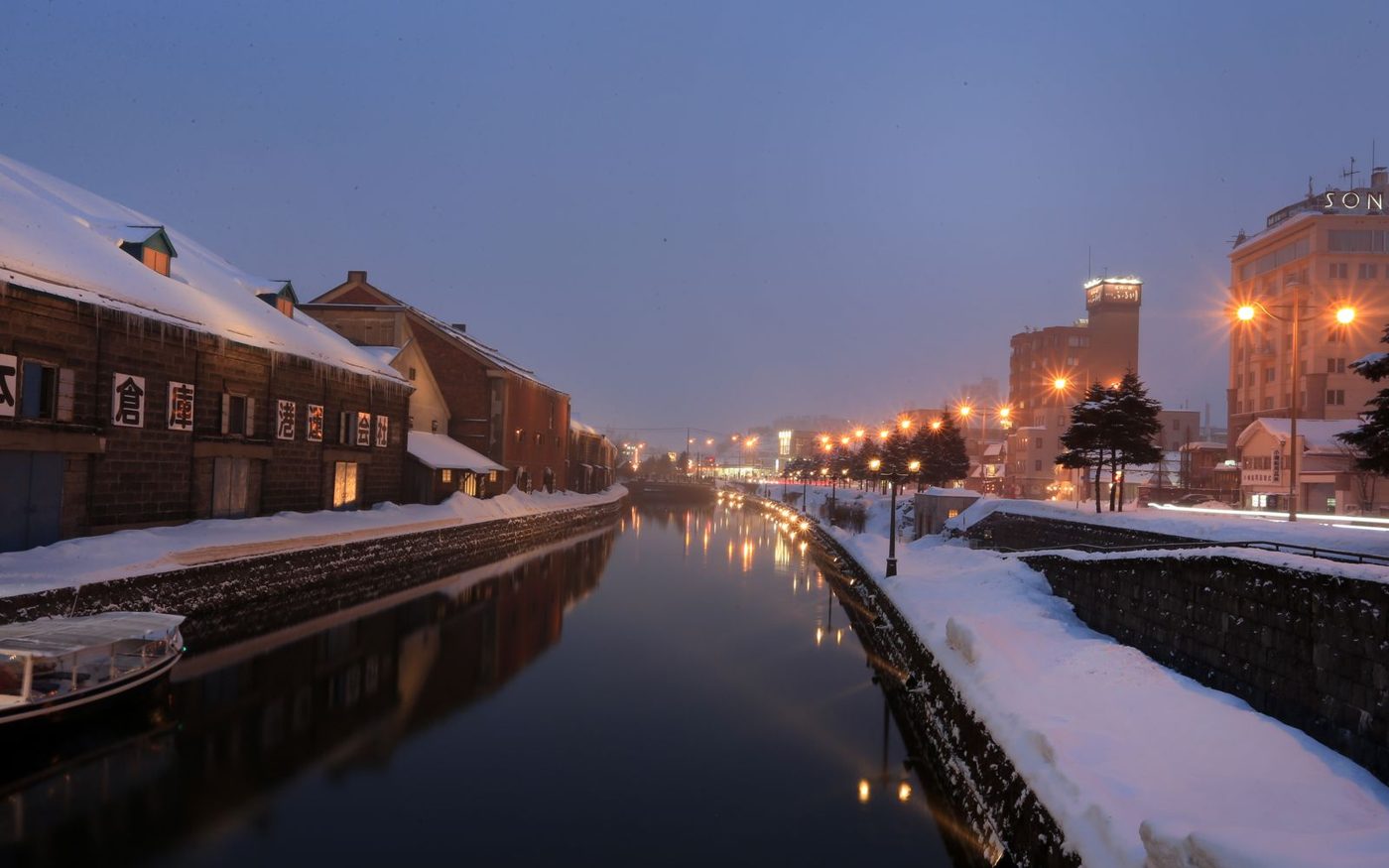 The Otaru canal at dusk in winter, snow on the banks and warehouse buildings lit along the water