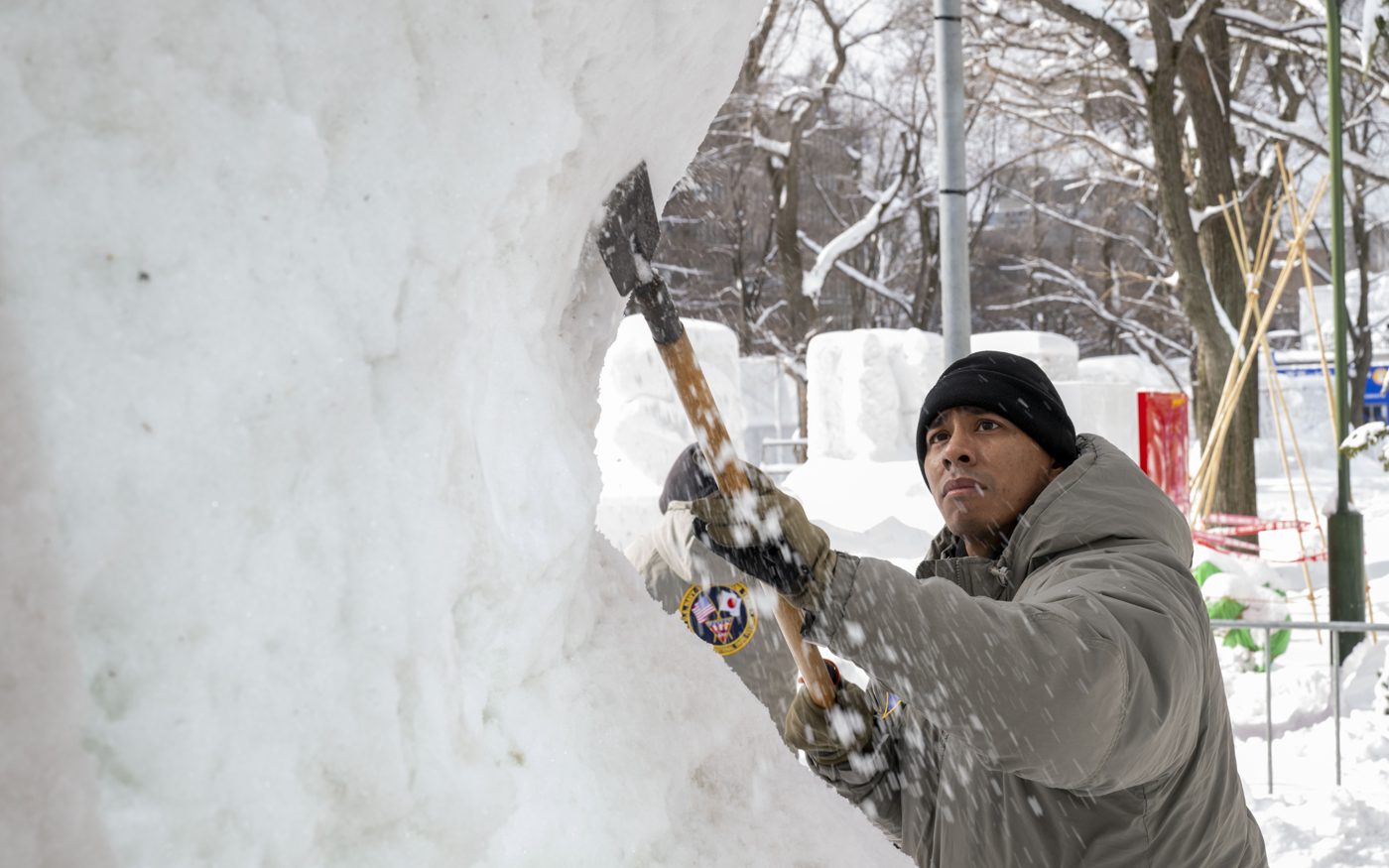 A worker swinging a long-handled tool at a tall block of packed snow, snow chips flying