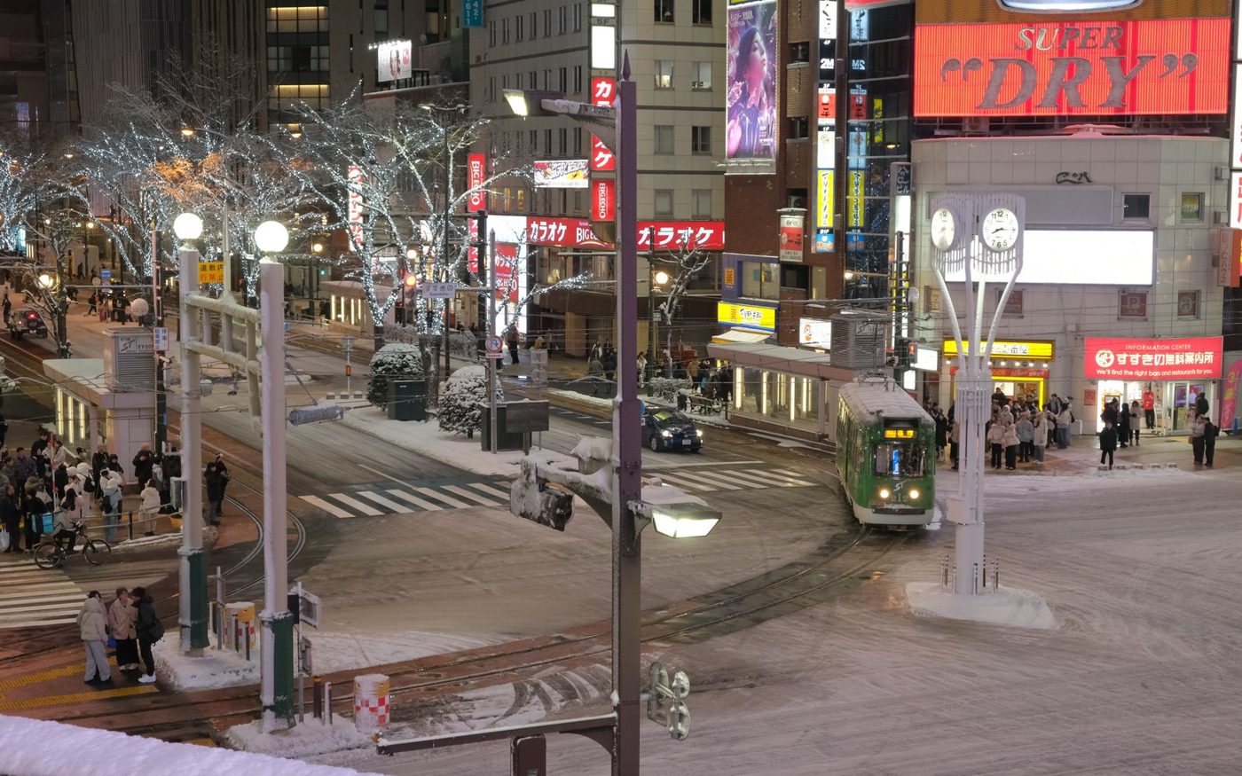 A snow-covered street at night with a tram, pedestrians and lit shop signs