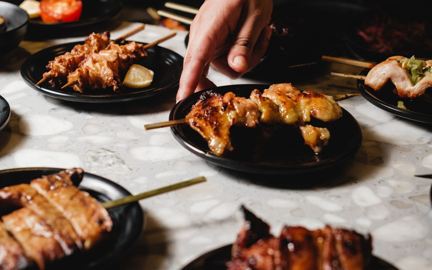 Several small black plates of grilled chicken skewers on a speckled stone table, one skewer being lifted