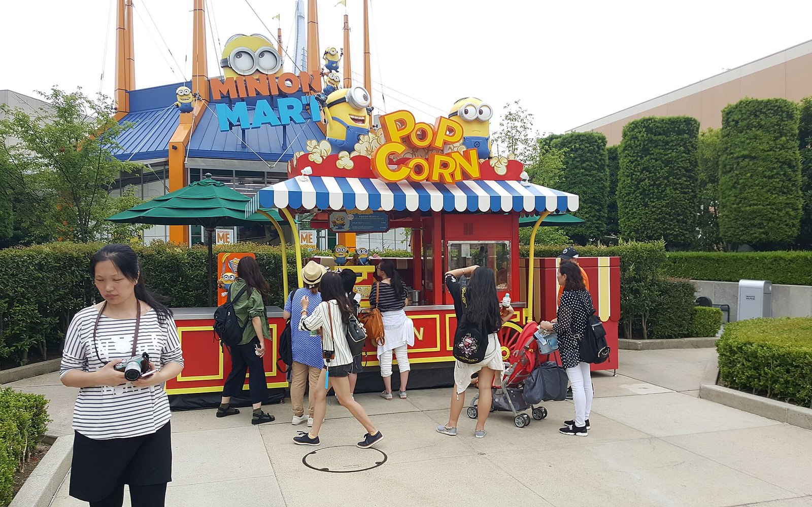 A Minion-themed Pop Corn stand with a striped awning, customers at the counter and the Minion Mart behind it