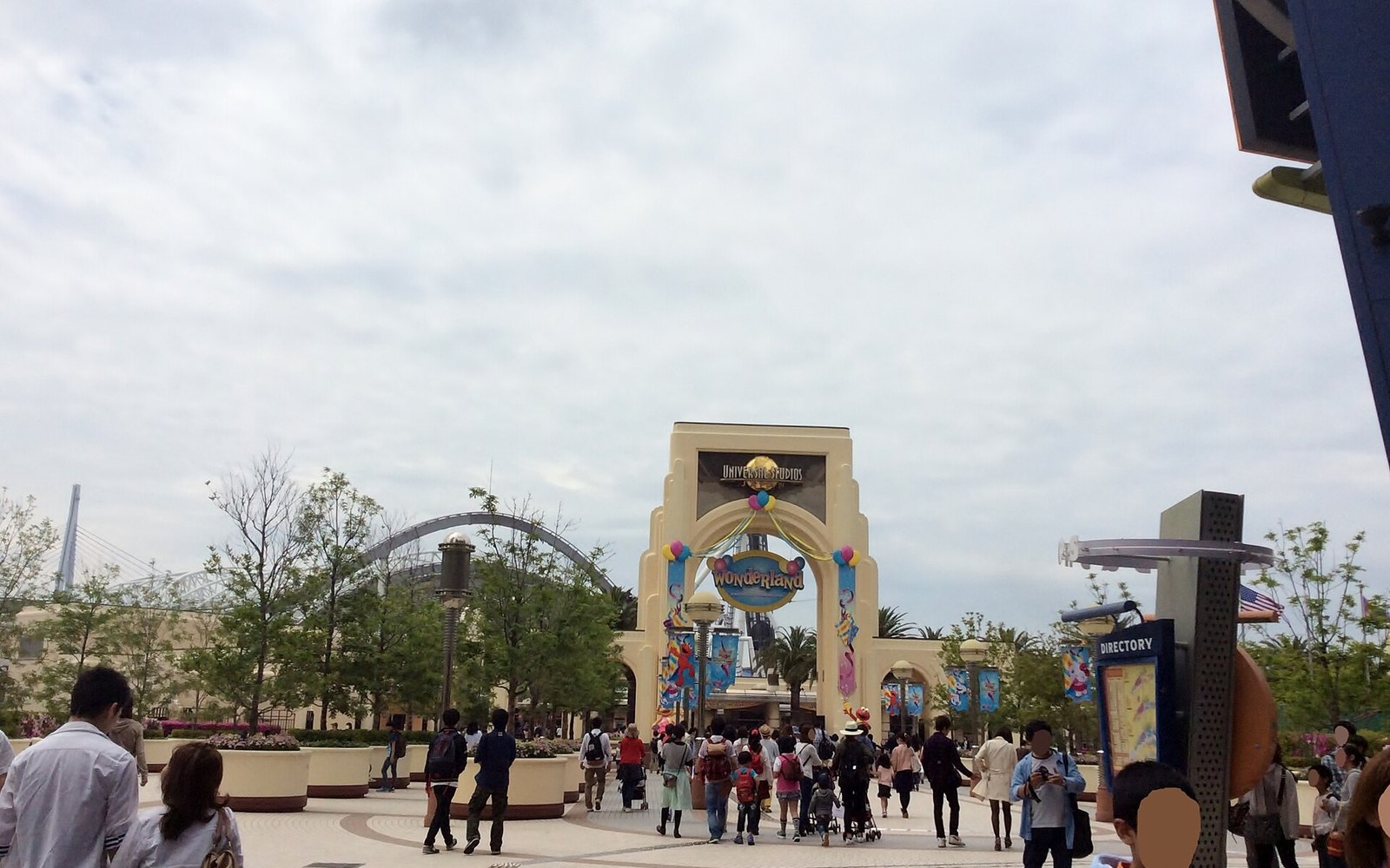Visitors crossing a paved plaza towards the Universal Wonderland entrance arch, with a roller coaster visible beyond it