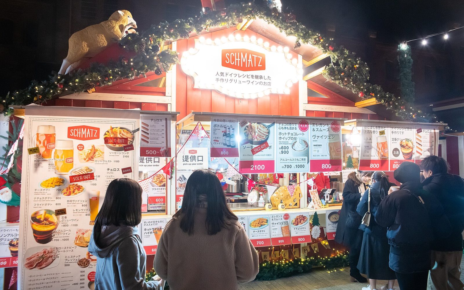 A red wooden market hut at night with menu boards lit up and customers queuing at the counter