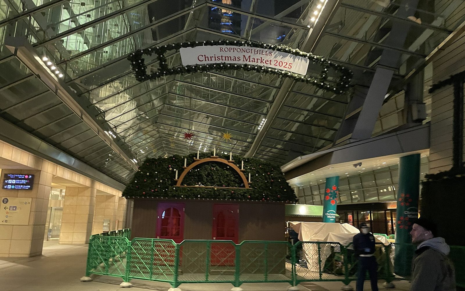 A wooden market hut framed with evergreen garland and red ornaments under a glass roof, with staff setting up