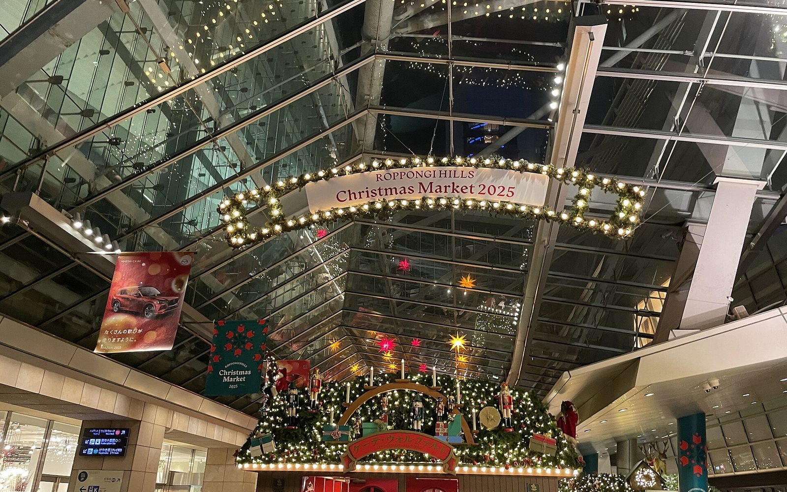 A dense evening crowd in front of a large Christmas hut covered in garland and lights, under a glass and steel roof