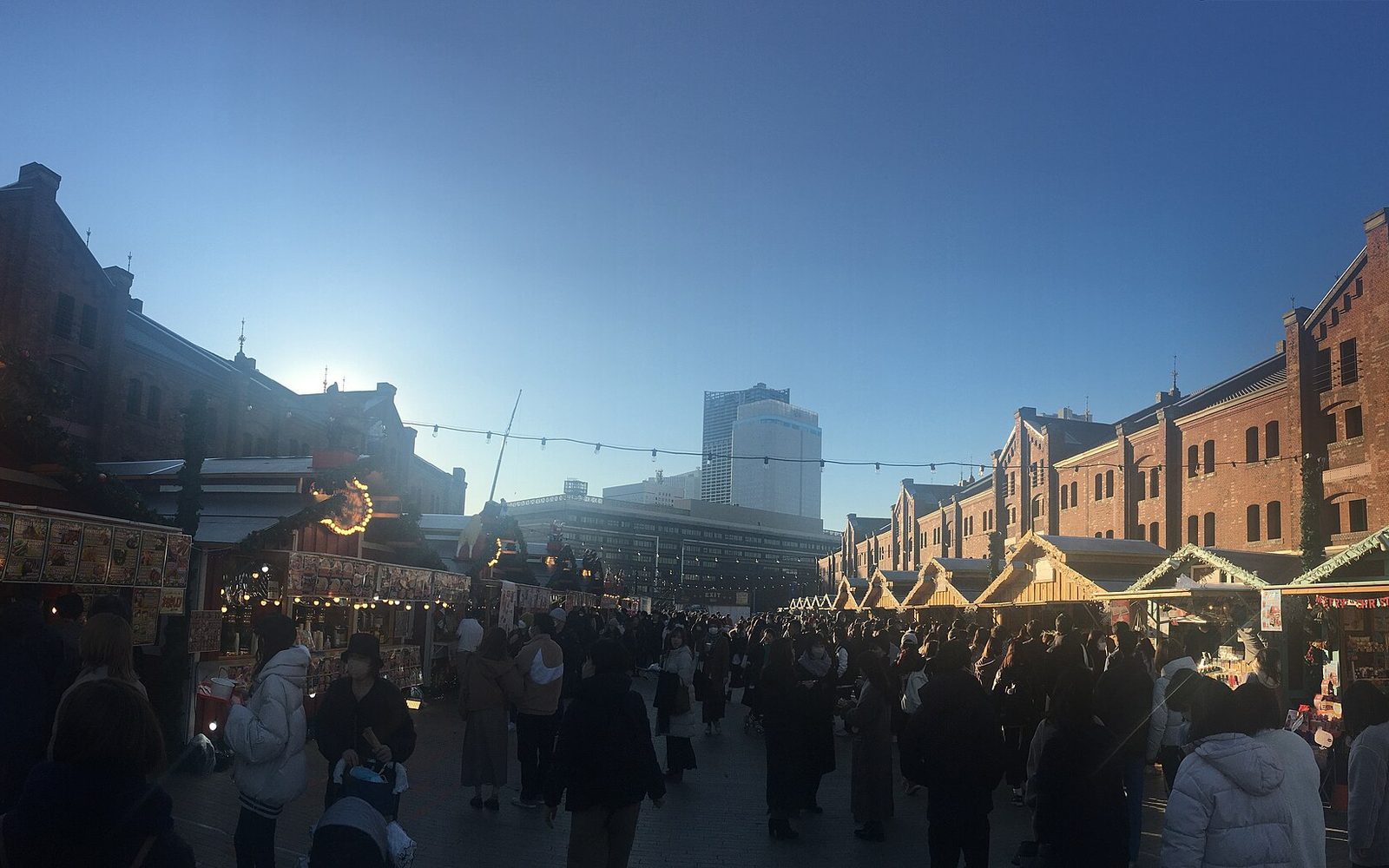 Wooden market huts with warm lights in front of the long red brick warehouse buildings, with a crowd in winter coats